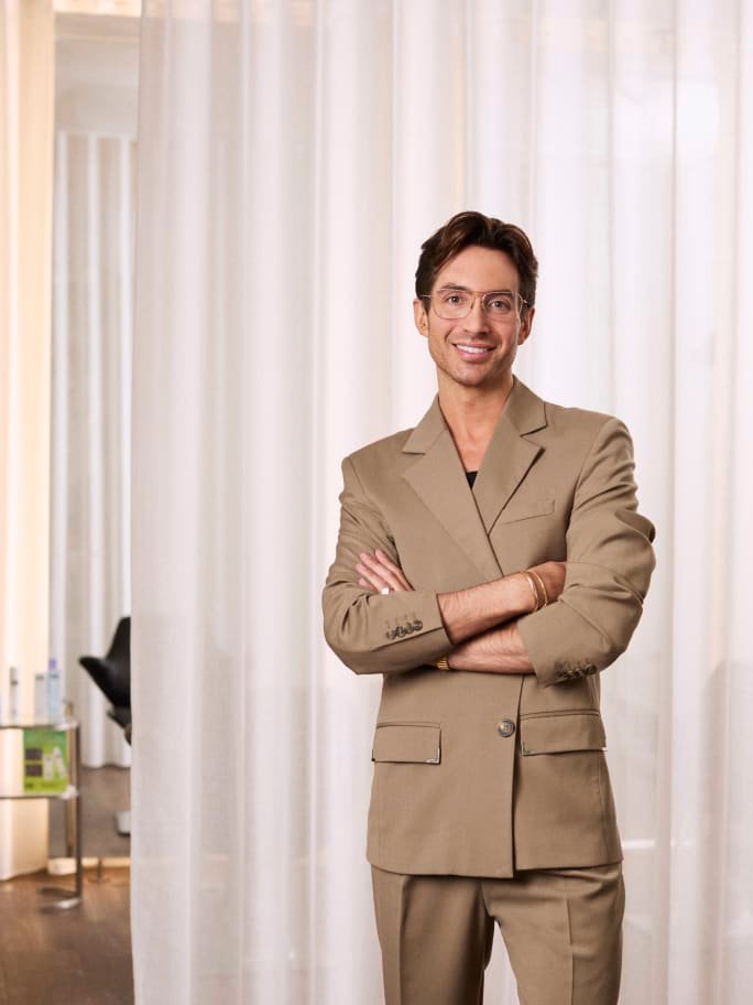 Portrait photo of a smiling man in a tan blazer suit with arms crossed, wearing glasses and gold jewelry, standing in front of sheer white curtains in a salon setting.