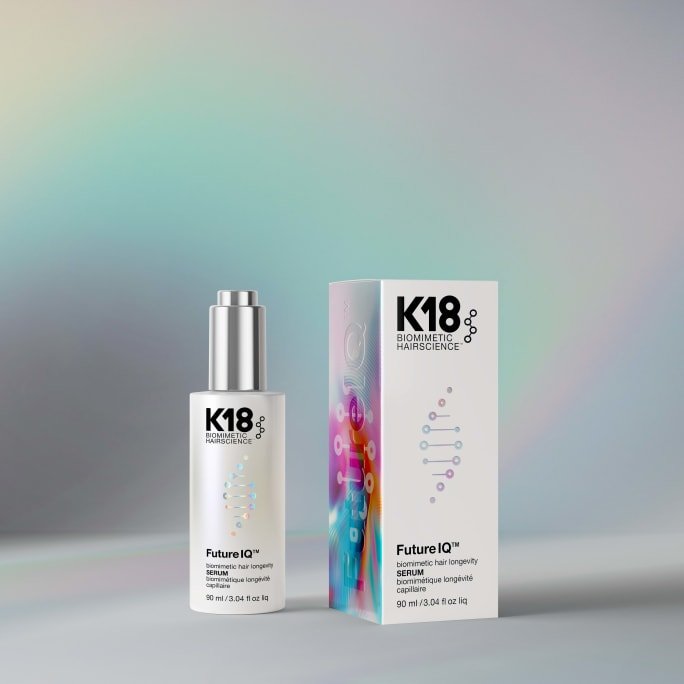 Product photo of a K18 Biomimetic Hairscience Future IQ hair longevity serum bottle (90 ml) standing beside its matching packaging box, both featuring a colorful DNA-strand graphic, set against a soft pastel-rainbow gradient background.