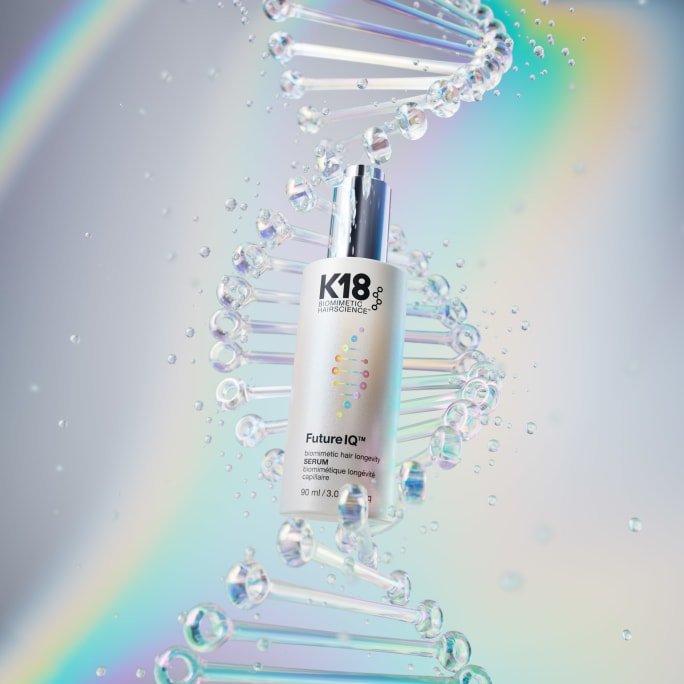 CGI product image of a K18 Biomimetic Hairscience Future IQ hair longevity serum bottle (90 ml) standing in front of a large glass-like DNA double-helix structure with water droplets, set against a soft iridescent rainbow background.