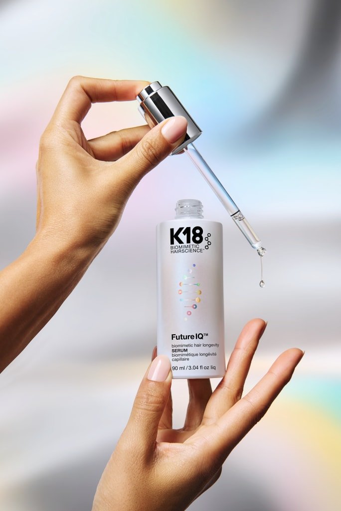 Product photo of a hand holding a K18 Biomimetic Hairscience Future IQ hair longevity serum dropper with a droplet of liquid falling, positioned above the open bottle (90 ml), against a soft pastel-rainbow blurred background.