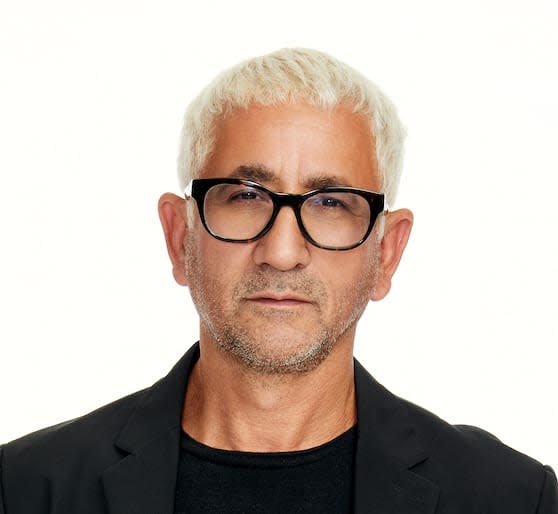 Professional headshot of a person with short platinum blond hair, black-framed glasses, and light facial stubble, wearing a black blazer over a black shirt against a plain white background, looking directly at the camera with a neutral expression.
