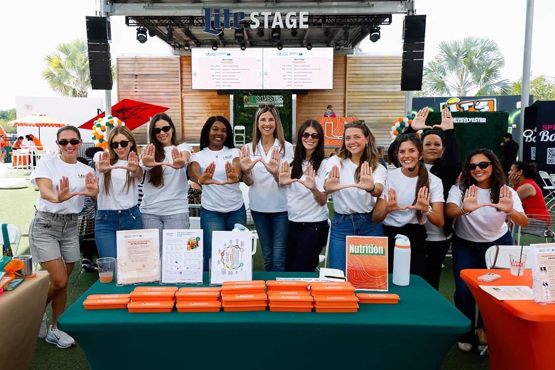 Staff wearing Sylvester-branded shirts stand behind an information table at the Sylvester Comprehensive Cancer Center survivorship celebration, holding their hands in a “U” hand sign, with educational materials and giveaway items displayed in front of a stage.