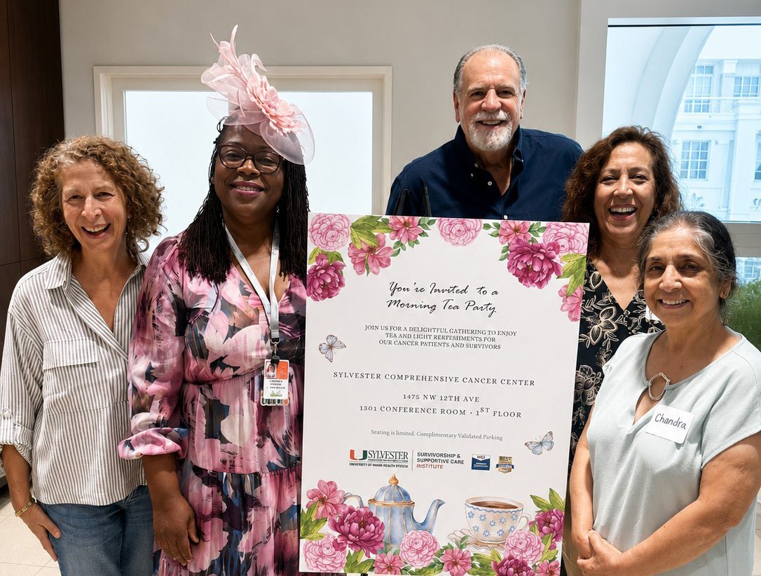 Group of five people standing indoors at Sylvester Comprehensive Cancer Center holding a large printed sign for a tea party event, decorated with pink floral illustrations and a teapot graphic. The sign includes event details about a morning tea party for cancer patients and survivors at Sylvester Comprehensive Cancer Center.