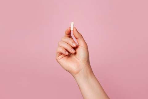 Hand holding small white pill against pink background.