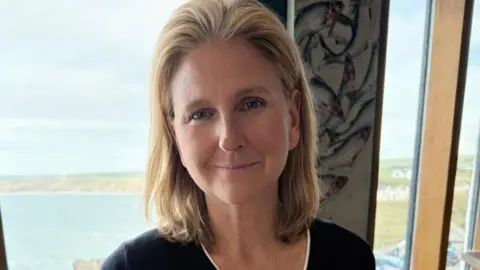 Laura Mayes Wiltshire councillor Laura Mayes with shoulder-length blonde hair and a black top with a sea-view through windows in the background.