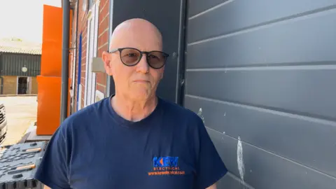 BBC A man with a bald head and black sunglasses wearing a navy blue t-shirt with the logo 'Kew Electrical' on it, looking straight-faced and standing beside a garage door with some gull excrement on it at the Hopton Industrial Estate in Devizes.