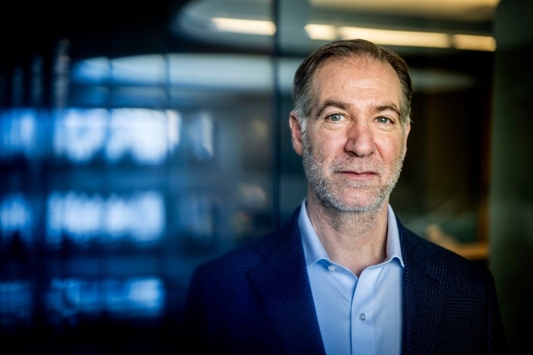 Portrait of Charles Hillman wearing a blazer over a blue shirt posing inside of a modern building.