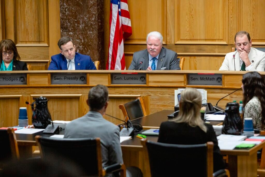 State. Sen. Jay Luneau, seated behind a dais with other lawmakers, asks a question to Surgeon General Dr. Evelyn Griffin, seated a table facing him.
