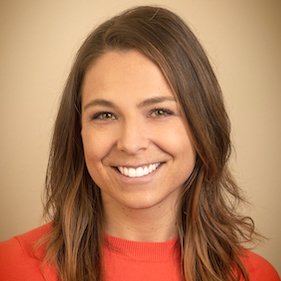 Close-up headshot of a person with long brown hair, smiling, wearing a coral top against a plain beige background.