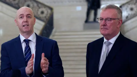 PA Media Jon Burrown and Mike Nesbitt. Jon os bald, wearing a white shirt and blue suit. Mike has short grey hair, wearing a white shirt, grey tie and black blazer.