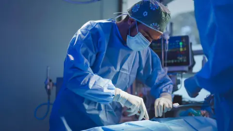 Getty Images A stock image of a surgeon in blue scrubs leaning over a patient holding tools.