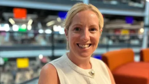 Michelle is smiling at the camera, she is wearing a cream sleeveless top and is sat on orange seats, with an open-plan, multi-level office setting in the background.