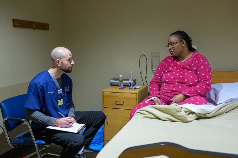 Male technologist in scrubs seated beside a patient in a pink robe on a hospital bed.