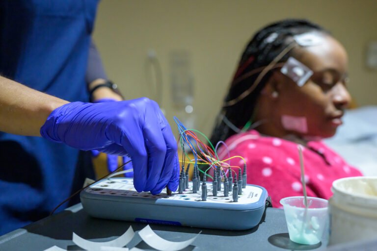 Gloved hands connecting more than a dozen colored electrode wires to a small transmission device.