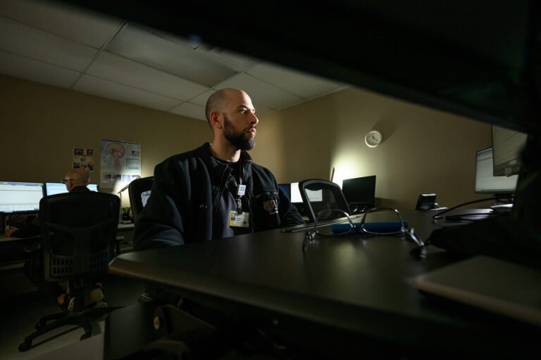 Bearded technologist seated at a computer in a dimly lit monitor room.