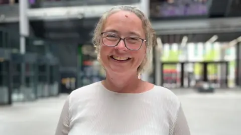 Rebeccah is wearing a white top and is smiling at the camera. She is stood in a large open office space.