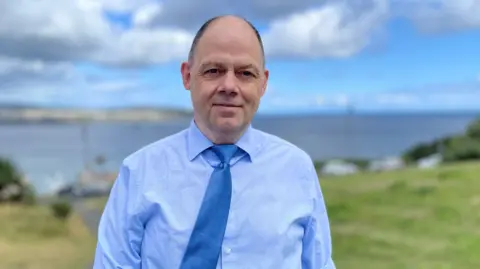 BBC Chris Stockport is mainly bald with short dark hair above his ears. He is wearing a light blue shirt with a darker blue tie and standing on grass with Douglas bay behind him. There are lots of clouds in the otherwise blue sky.