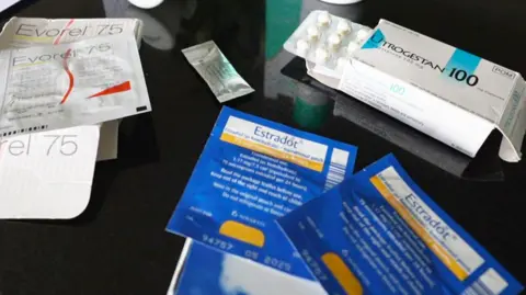 BBC Different types of HRT are laid out on a table, including tablets, patches, spray and gel. In the centre is a small sachet of testosterone gel.