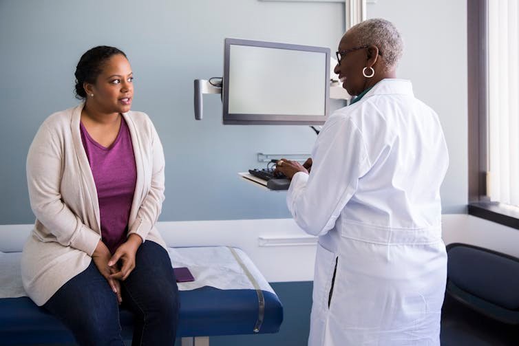 Patient sitting on exam room table talking to doctor, who is standing up at computer